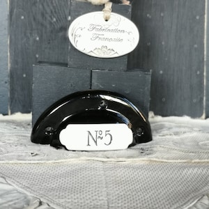 Könnte beinhalten: Schwarze Keramik-Hausnummer-Plakette mit weißem Hintergrund und der Nummer "N° 5" in silbernen Lettern. Die Plakette ist an einer schwarzen Halbkreisbasis befestigt. Ein silbernes ovales Schild mit dem Text "Fabrication Française" ist im Hintergrund sichtbar.