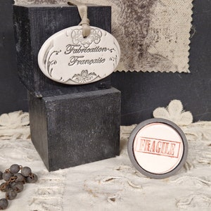 May include: A white oval ceramic tag with the text "Fabrication Française" on a black background. A black and white fabric swatch is in the background. A black and white beaded necklace is in the foreground. A round, black and white sticker with the word "FRAGILE" in red letters is on a white surface.