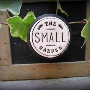 May include: A white ceramic sign with black lettering that reads "The Small Garden". The sign is attached to a wooden frame with a green ivy plant growing around it.