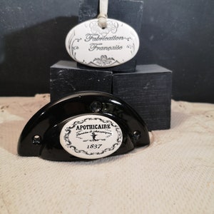 May include: A black ceramic cabinet knob with a white porcelain insert. The insert features the text "Apothecaire" with a decorative design and the year "1837". A separate white porcelain oval tag with the text "Fabrication Française" is also shown.