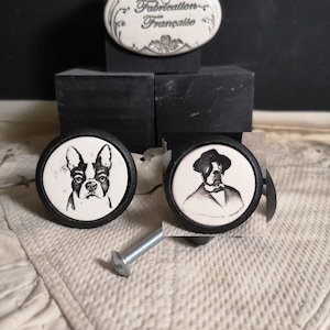 May include: Two ceramic drawer knobs with black trim. One knob features a black and white illustration of a Boston Terrier dog. The other knob features a black and white illustration of a man wearing a hat and a suit. The knobs are on a white surface with a black screw.