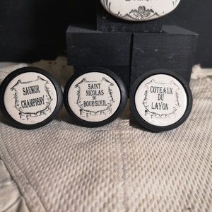 May include: Three white ceramic bottle stoppers with black lettering. The stoppers are round and have a raised design. The text on the stoppers reads "Saumur Champigny", "Saint Nicolas de Bourgueil", and "Coteaux du Layon".
