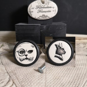 May include: Two black and white ceramic knobs with a cat design. One knob features a cat with a monocle and the other features a cat wearing a top hat. The knobs are on a black surface with a white oval sign that reads "Fabrication Française".