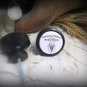 以下が含まれることがあります： 黒いキャビネットノブが2つ。1つのノブには、黒い文字で「Fabrique de Cravates - M. Mathieu.」と書かれた白い円形のインサートがあります。インサートには、スーツとネクタイを着た男性の白黒のイラストが描かれています。