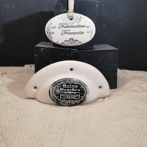 Peut inclure: Accessoire de salle de bain en céramique blanche avec une étiquette ovale noir et blanc qui indique "Bains Douches" et "Specialite de Bains et Douches". L'accessoire a une petite étiquette ovale qui indique "Fabrication Française".