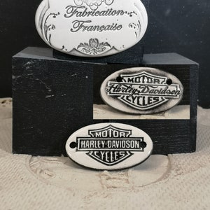 Peut inclure: Deux étiquettes ovales en céramique blanche. L'étiquette du haut porte le texte "Fabrication Française" dans une police d'écriture décorative. L'étiquette du bas porte le logo Harley Davidson en noir avec le texte "Motor Cycles".