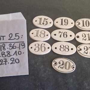 Puede incluir: Un sobre blanco con el texto "Lote 25: 15.18.36.19 76.88.10. 21.27.20" escrito en él. Hay diez etiquetas ovaladas de cerámica blanca con números negros en ellas. Los números en las etiquetas son 15, 19, 10, 18, 76, 21, 36, 88, 27 y 20.
