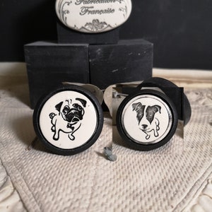 Peut inclure: Deux boutons de tiroir en céramique avec des bases noires et des dessus blancs. Le bouton de gauche présente une illustration de carlin noir et blanc. Le bouton de droite présente une illustration de Jack Russell Terrier noir et blanc. Les deux boutons portent le texte "Fabrication Française" au dos.