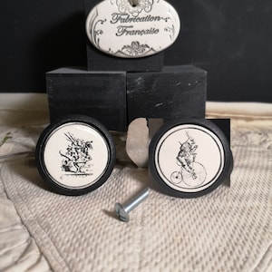 May include: Two black ceramic knobs with white illustrations. One knob features a whimsical illustration of a person riding a unicycle, the other features a person holding a teacup and teapot. The knobs are displayed on black blocks with a white oval tag that reads "Fabrication Française".
