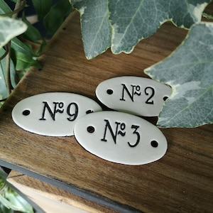 以下が含まれることがあります： 黒い文字で書かれた白いセラミック製の楕円形のタグが3つ。タグには「No. 2」、「No. 9」、「No. 3」と番号が付けられています。