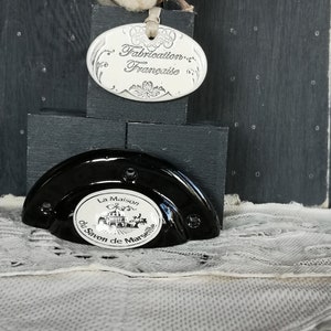 Könnte beinhalten: Eine schwarze Keramik-Seifenschale mit einem weißen ovalen Etikett, auf dem "La Maison du Savon de Marseille" steht. Die Seifenschale steht auf einer weißen Spitzenoberfläche. Die Seifenschale ist mit einem kleinen weißen Keramikhaus mit Schornstein und Fenster verziert. Die Seifenschale ist auch mit einem kleinen weißen Keramikbaum verziert. Die Seifenschale steht auf einer schwarzen Holzoberfläche. Oberhalb der Seifenschale befindet sich ein weißes ovales Etikett mit der Aufschrift "Fabrication Française".