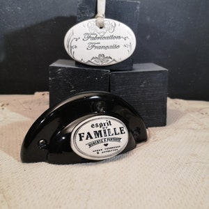Puede incluir: Pomo de puerta de cerámica negra con una etiqueta ovalada de cerámica blanca que dice "esprit de FAMILLE MOMENTS A PARTAGER AMOUR TENDRESSE & AFFECTION". El pomo está sobre una superficie negra con una etiqueta ovalada de cerámica blanca que dice "Fabrication Française".