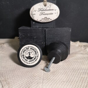 Könnte beinhalten: Schwarzer Keramik-Schrankknopf mit einem weißen runden Design mit dem Text "Rio de Janeiro" und einem stilisierten Bild einer Person, die am Strand steht. Der Knopf ist an einer silbernen Schraube befestigt. Ein weißes ovales Schild aus Keramik mit schwarzem Text lautet "Fabrication Française".