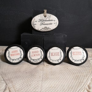 May include: Five white ceramic knobs with black trim and red lettering. The knobs are labeled with French wine regions: Auxey-Duresses, Aloxe-Corton, Beaune, Bouzeron. A white ceramic oval tag with black lettering reads "Fabrication Française".