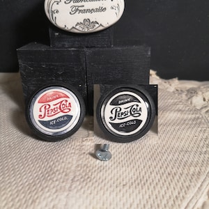 May include: Two black and white Pepsi-Cola bottle caps with the text "Drink Pepsi-Cola Ice Cold" on each. The bottle caps are on a black surface. A silver screw is in the center of the image.