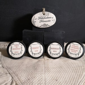 May include: Five white ceramic bottle stoppers with black lettering. Each stopper features a different French wine region name and vintage year. The stoppers are arranged on a white fabric surface. The text on the stoppers reads: "Cote de Bergerac 2005", "Pechredon 1995", "Saussignac 2009", "Chateau Laurens, Margillac Valley".