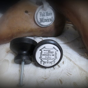 May include: Two black ceramic cabinet knobs with a silver screw. The knob on the right has a white porcelain insert with black text that reads "Compagnie Colombiere Specialite Chocolat & The".