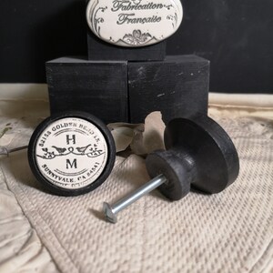 Könnte beinhalten: Schwarzer Keramik-Knopf mit einem weißen Keramikeinsatz mit dem Text "Grass Golden Bend Ln Sunnyvale CA 94087" und den Buchstaben "H M". Der Knopf ist an einer silbernen Schraube befestigt. Im Hintergrund ist eine weiße ovale Keramikplakette mit dem Text "Fabrication Française" zu sehen.