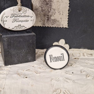 May include: A white ceramic tag with the text "Fabrication Française" and a black and white round tag with the text "Fenouil" on a white lace tablecloth.