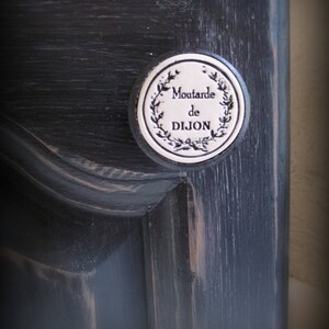 May include: A black wooden cabinet door with a white ceramic knob. The knob has a black and white design with the text "Moutarde de Dijon".