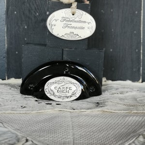 May include: Black ceramic half-dome with a white inscription that reads "Carpe Diem" and a decorative floral design. The half-dome is sitting on a black wooden base with a white oval ceramic tag that reads "Fabrication Française".