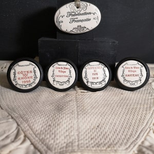 May include: Five white ceramic bottle stoppers with black trim and red text. The stoppers feature different French wine regions and years. The text on the stoppers reads: "Cotes du Rhone 1998", "Cotes du Rhone Villages Vacqueyras 1967", "Chateauneuf du Pape 2012", "Cotes du Rhone Rasteau 1944".