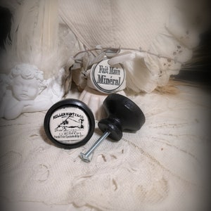 May include: Two black cabinet knobs with a vintage aesthetic. One knob features a circular design with the text "Keller Kurtz & Co." and a graphic. The other knob is detached, showing the screw. The background includes a white lace fabric and a small angel statue.