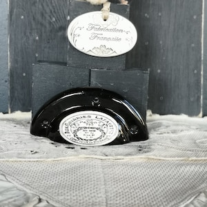 Peut inclure: Un bouchon de bouteille en céramique noir et blanc avec le texte "Aux Galeries Lafayette, Patron 763, Tête Intérieur N° 32, Comptoir de Paris" en cercle autour du bord. Le bouchon est posé sur un bloc de bois noir et une dentelle blanche.