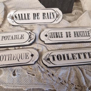 May include: Five white ceramic signs with black lettering. The signs read "Salle de Bain", "Eau Potable", "Meuble de Famille", "Bibliotheque", and "Toilettes".