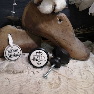 May include: Two ceramic knobs with black bases. One knob features the word "BISTRO" in black lettering on a white background. The other knob has the text "Fait Main Mineral" in black lettering on a white background.