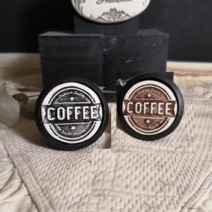 May include: Two round ceramic knobs with a black background and white lettering that says "Premium Quality Coffee Freshly Roasted". One knob has a brown coffee bean design and the other has a black coffee bean design.
