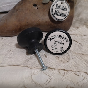 May include: Two black cabinet knobs with a screw and a decorative ceramic face. One knob features the word "Bibliothèque" with a stack of books. The other knob is plain black. The knobs are on a lace surface.