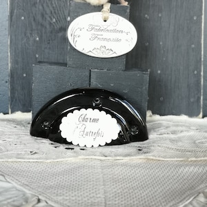Peut inclure: Une poignée de tiroir noire semi-circulaire avec le texte "Charme d'Autrefois" sur une étiquette blanche. Au-dessus, une étiquette indique "Fabrication Française". L'image comprend une boule de coton et est posée sur un fond gris foncé.