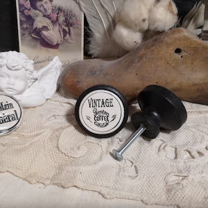 May include: Two black and white ceramic knobs with a vintage coffee design and the text "Vintage Premium Coffee Collection". One knob is attached to a black metal screw. The knobs are on a white lace surface.