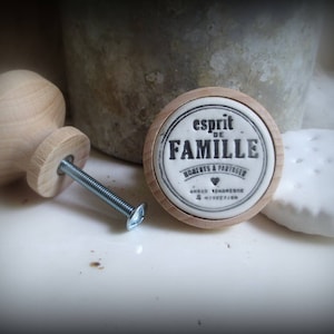 Może przedstawiać: Drewniany uchwyt do szafki ze srebrną śrubą obok okrągłego drewnianego i ceramicznego uchwytu z napisem "esprit DE FAMILLE". Na zdjęciu znajduje się również biały ceramiczny dysk.