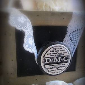 Puede incluir: Una bobina de hilo de bordar DMC en blanco y negro con el texto "Cotton Silk and Flax over 500 Colours DMC". La bobina está sobre una superficie de madera con encaje blanco.