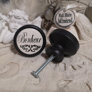May include: Two black ceramic cabinet knobs with a silver screw. One knob has a white ceramic insert with the word "Bonheur" in black script and a decorative design. The other knob is plain black.