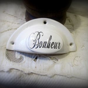 Puede incluir: Tirador de cajón de cerámica blanca con forma redondeada y un óvalo en relieve en el centro. El óvalo tiene la palabra "Bonheur" escrita en cursiva negra.