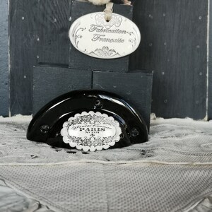 Może przedstawiać: Czarny ceramiczny korek do butelki z białą porcelanową etykietą z napisem "Jante-Cavenne PARIS N°217". Korek leży na białej koronkowej serwetce. Biała porcelanowa owalna etykieta z czarnym tekstem głosi "Fabrication Française".