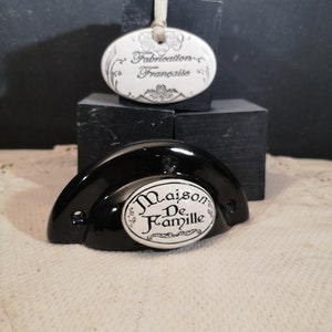 Könnte beinhalten: Ein schwarzer Keramik-Schrankknauf mit einem weißen Porzellan-Einsatz. Der Einsatz trägt die Worte "Maison De Famille" in einer dekorativen Schrift.