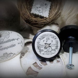 May include: Two white ceramic drawer knobs with black bases. The knobs feature a black and white design with the text "LINGERIE - PARIS - SOIETE" and a stylized image of a blacksmith's hammer.