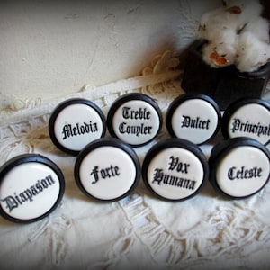 Peut inclure: Neuf boutons ronds noirs et blancs avec différents noms de registres d'orgue écrits en écriture noire. Les noms sont : Diapason, Forte, Vox Humana, Celeste, Melodia, Treble Coupler, Dulcet, Principal.