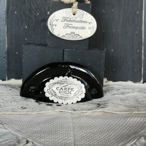 以下が含まれることがあります： 黒いセラミック製のドアノブに、白い磁器製のプレートが取り付けられており、「カルペ・ディエム」と書いてあります。ノブは黒い木製の台座の上に置かれており、白い磁器製のタグには「Fabrication Française」と書いてあります。