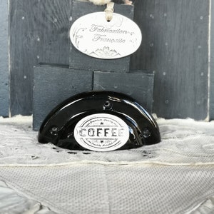 May include: Black ceramic coffee cup holder with a white label that says "Premium Quality Coffee Freshly Roasted". The holder is sitting on a white lace doily. There is a silver oval tag that says "Fabrication Française" on a black wooden block behind the holder.