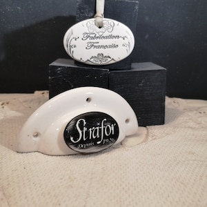 Peut inclure: Poignée de porte en céramique blanche avec une étiquette ovale noire qui indique "Strafor Depuis 1926". Une étiquette ovale en céramique blanche avec le texte "Fabrication Française" est suspendue au-dessus de la poignée.