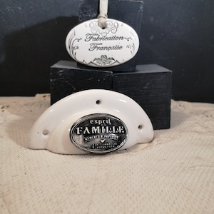Peut inclure: Deux pièces décoratives en céramique blanche. L'une est un pendentif ovale avec le texte "Fabrication Française" et un ruban. L'autre est une poignée de tiroir en forme de demi-lune avec le texte "esprit de FAMILLE".