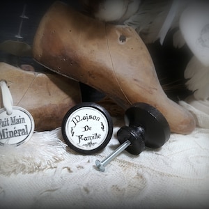 May include: Two black and white ceramic knobs with a screw. One knob has the text "Maison De Famille" on it. The other knob has a black base and a white top.