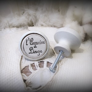 May include: Two white ceramic cabinet knobs. One knob has the text "L'Armoire à Linge" in black script. The knobs are on a white textured surface, with a metal screw attached. The knobs are likely for a linen cabinet.