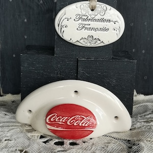 Peut inclure: Un bouchon de bouteille Coca-Cola en céramique blanche avec un logo Coca-Cola rouge. Le bouchon est en forme de demi-cercle et comporte deux trous sur le dessus. Le bouchon est posé sur une nappe en dentelle blanche. Une étiquette ovale en céramique blanche avec le texte "Fabrication Française" est en arrière-plan.