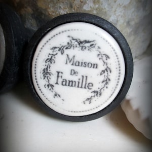 May include: A black and white ceramic knob with a black rim. The knob has a white background with black text that reads "Maison De Famille".